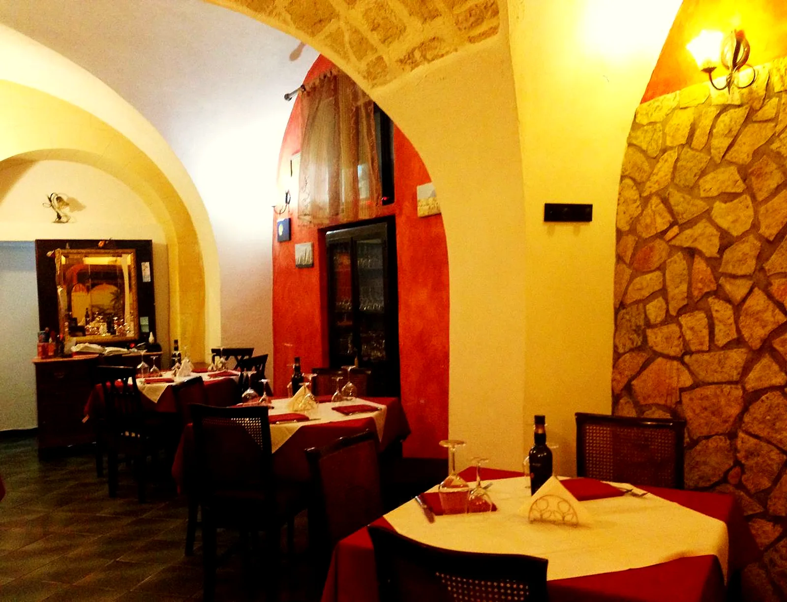 La parete in pietra a secco illuminata da un'applique, lo specchio dorato e i tavoli in fondo alla sala