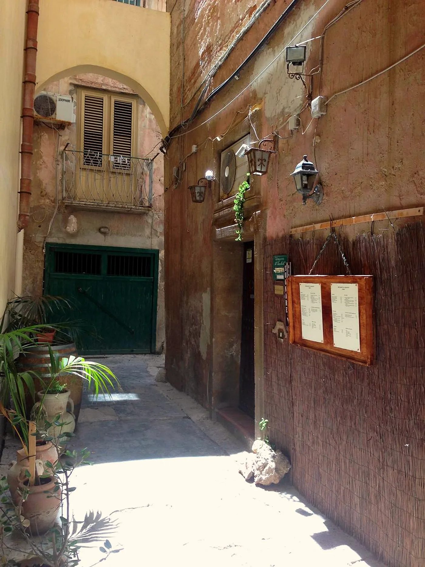 Il vicolo di giorno: la porta scura del ristorante, i lampioni in ferro e la bacheca del menu appesa al canniccio